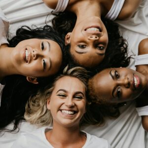 Four diverse women lying on a bed smiling at the camera, sharing a joyful moment.